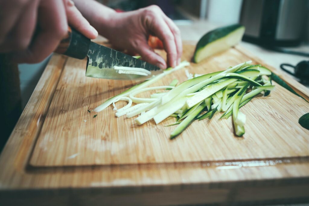 A person holding a stainless steel knife to thinly slice a vegetable on top of a brown wooden chopping board