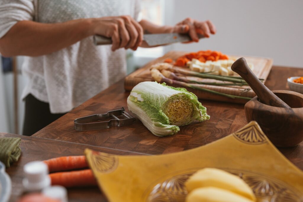 A person wearing a white shirt is holding a stainless steel knife to cut fresh vegetables on top of a brown wooden chopping board