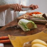 A person wearing a white shirt is holding a stainless steel knife to cut fresh vegetables on top of a brown wooden chopping board