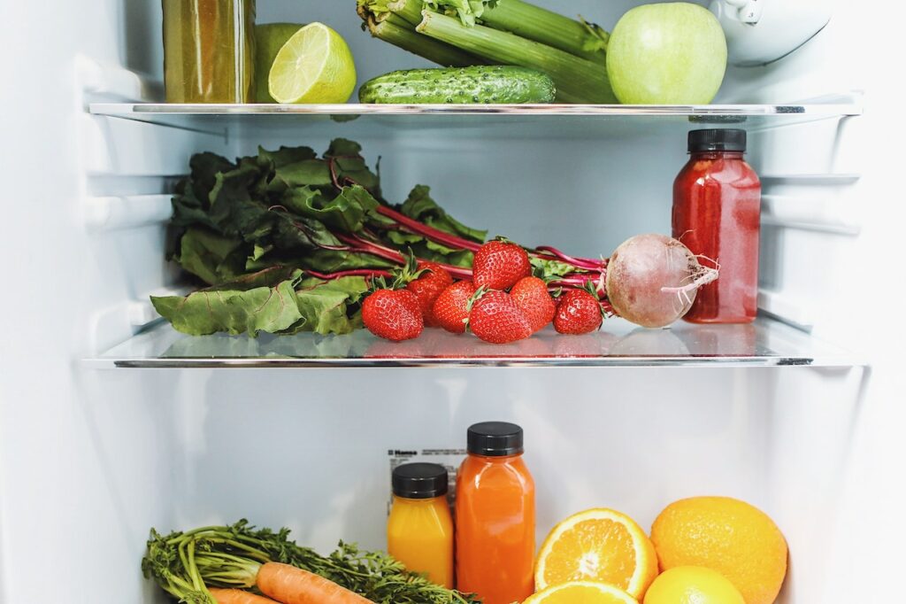 Fresh strawberries, sliced oranges, a turnip, carrots, juice in a clear bottle, and fresh vegetables inside a white fridge