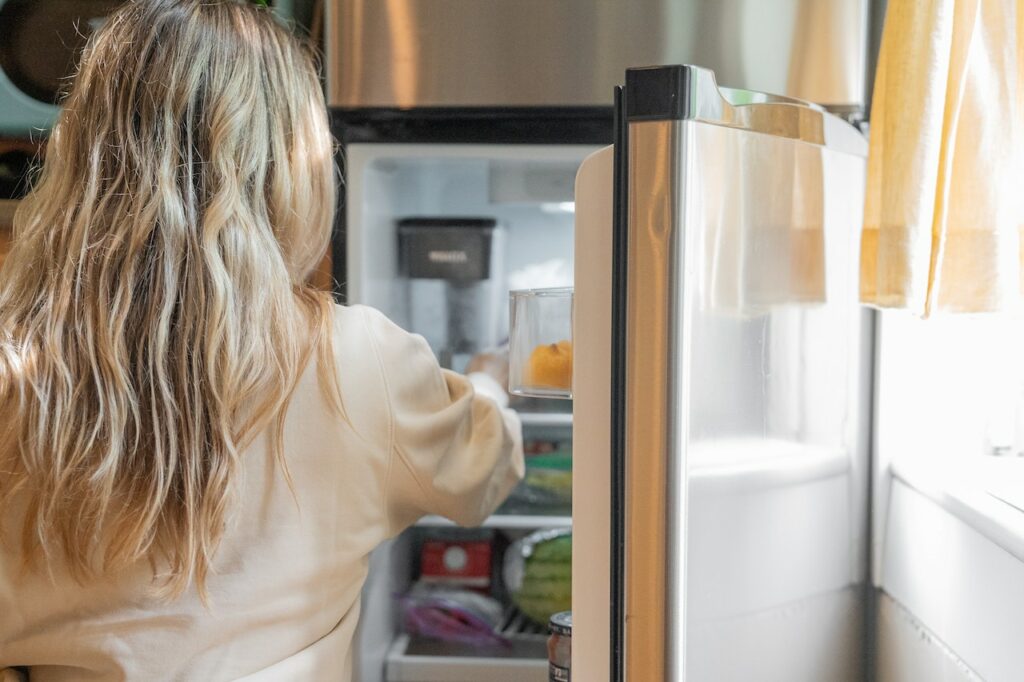 A woman wearing cream colored long sleeves is putting food inside a white fridge near a yellow curtain