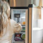 A woman wearing cream colored long sleeves is putting food inside a white fridge near a yellow curtain
