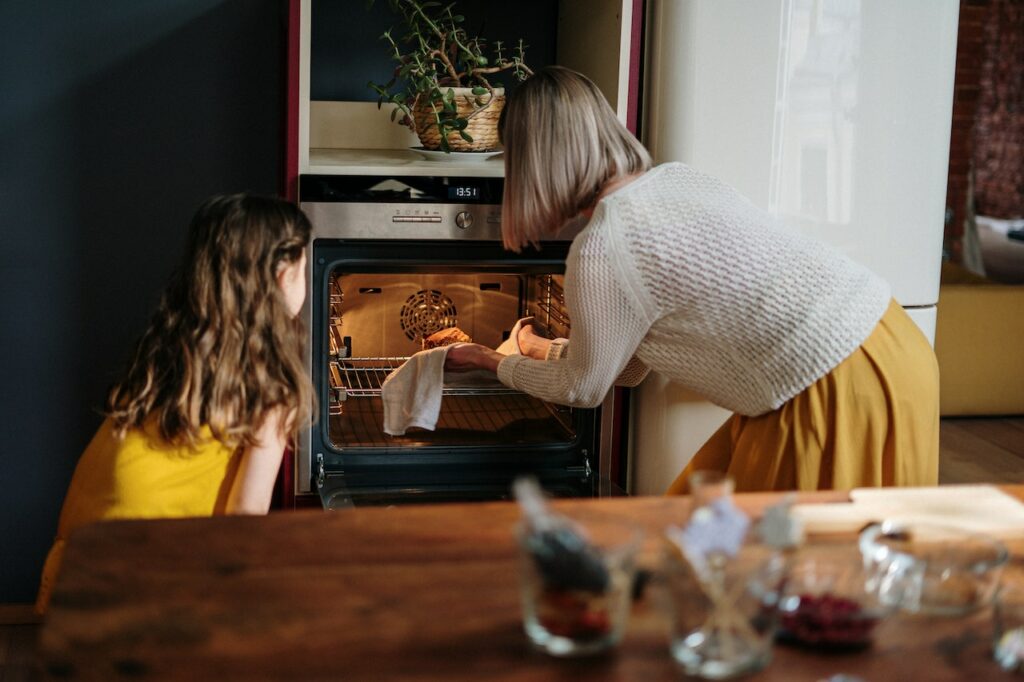 A woman in a white knitted sweatshirt and yellow skirt cooking in the oven with her daughter
