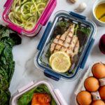 Clear food containers near a red onion, fresh eggs, oil, and vegetables on top of a marbled table