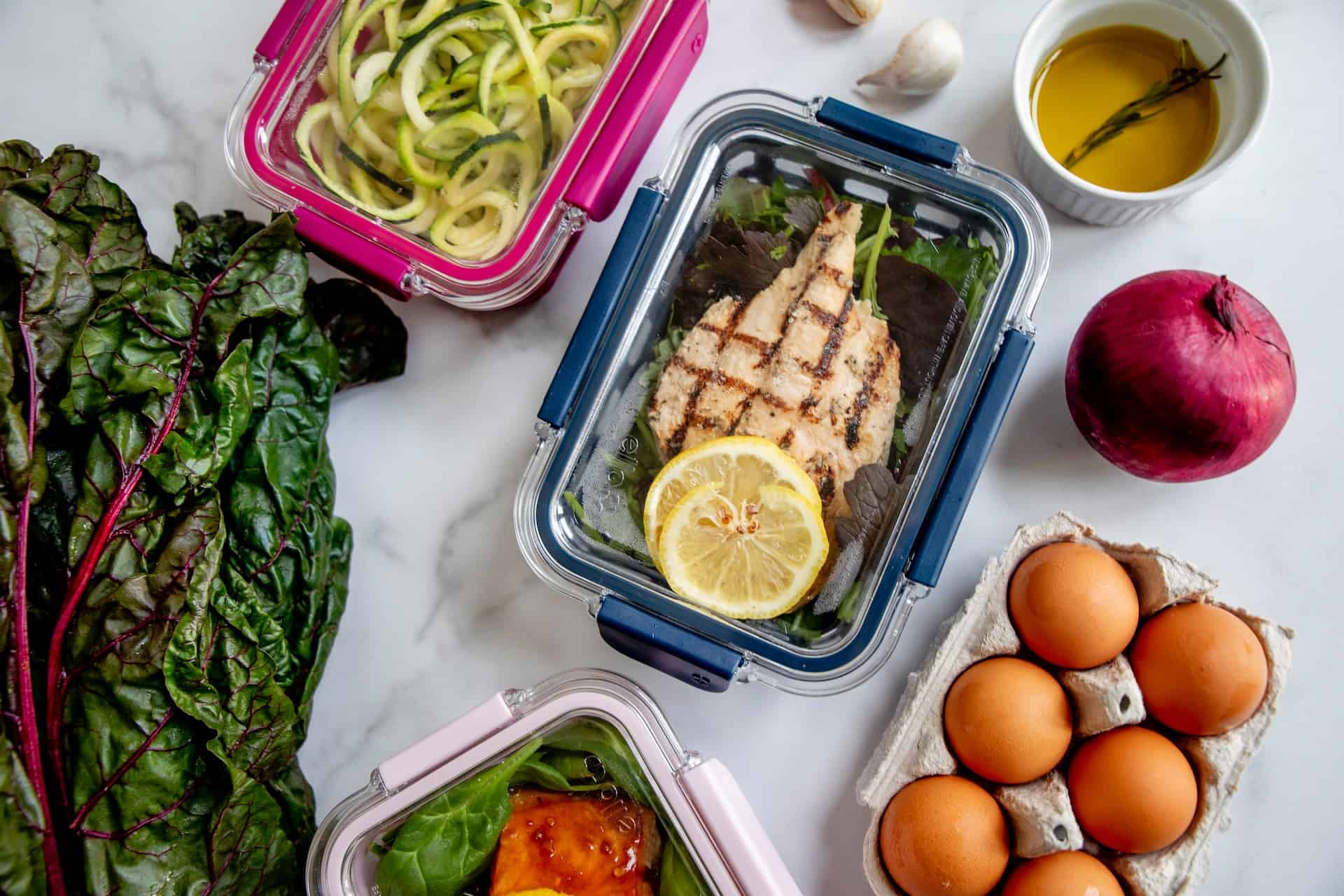 Clear food containers near a red onion, fresh eggs, oil, and vegetables on top of a marbled table