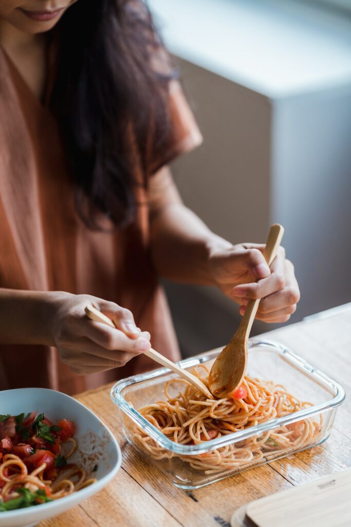 A woman wearing a brown oversized shirt is holding a wooden spoon and fork while placing pasta on a glass container on top of a brown wooden table