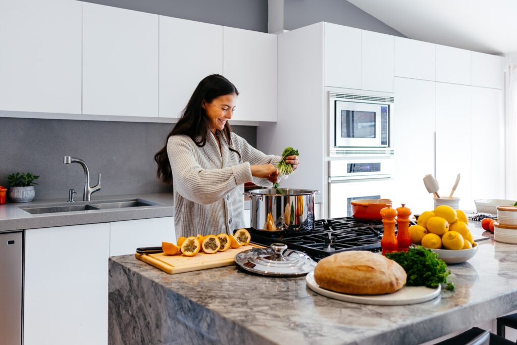 A woman wearing a gray sweater is putting vegetables inside a stainless steel pot on top of a black stovetop