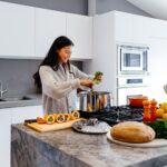 A woman wearing a gray sweater is putting vegetables inside a stainless steel pot on top of a black stovetop
