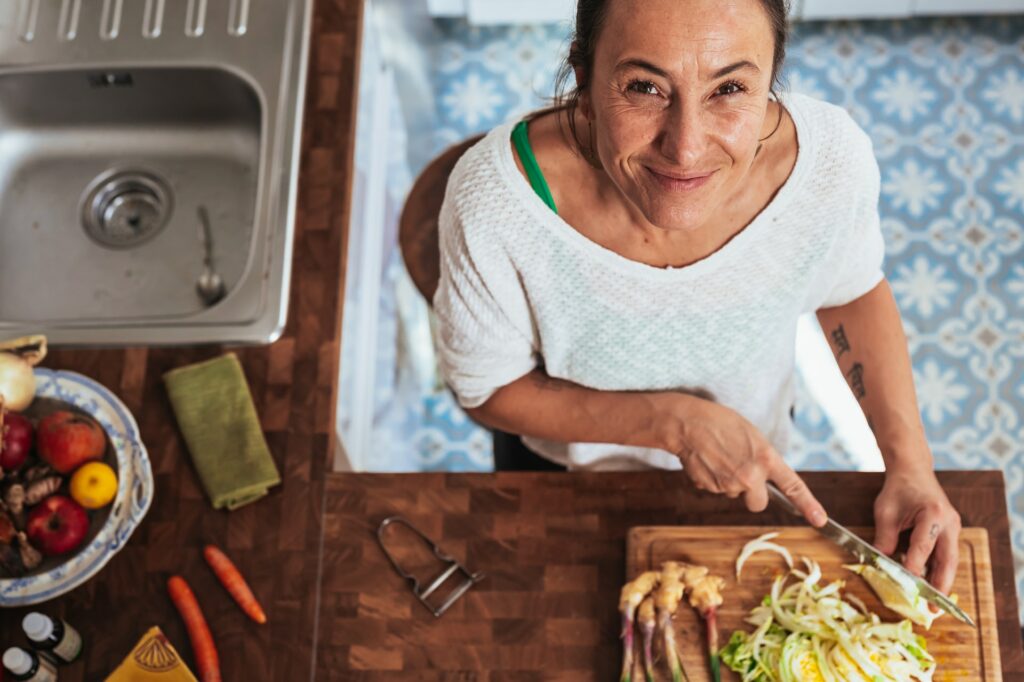 A woman wearing a white long-sleeve shirt is chopping vegetables using a stainless steel knife on a brown wooden chopping board
