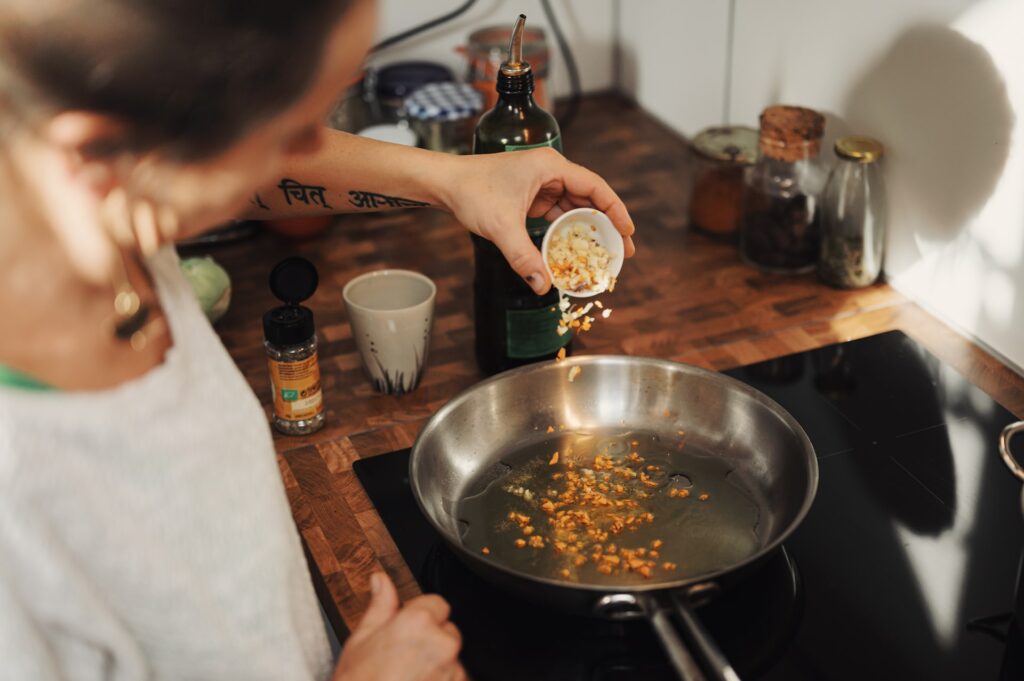 A woman wearing a gray shirt is putting minced garlic on a stainless steel pan that is placed on top of a black electric stove