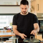 A man wearing a black shirt is cooking using a black pot with a glass cover on a black electric stove