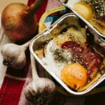 Egg in an aluminum pan near onion and garlic on top of a colorful pot holder on a brown wooden table