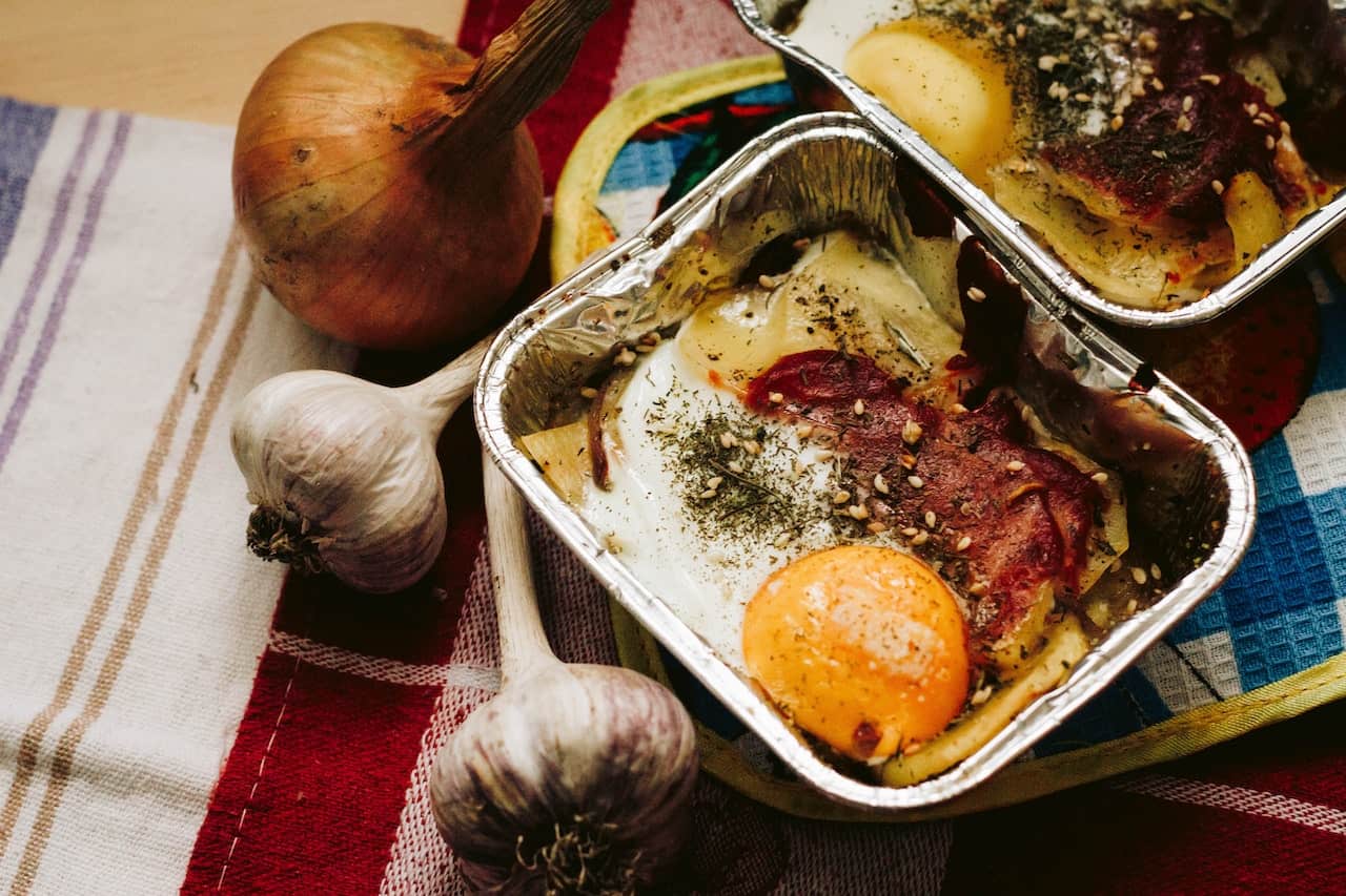 Egg in an aluminum pan near onion and garlic on top of a colorful pot holder on a brown wooden table