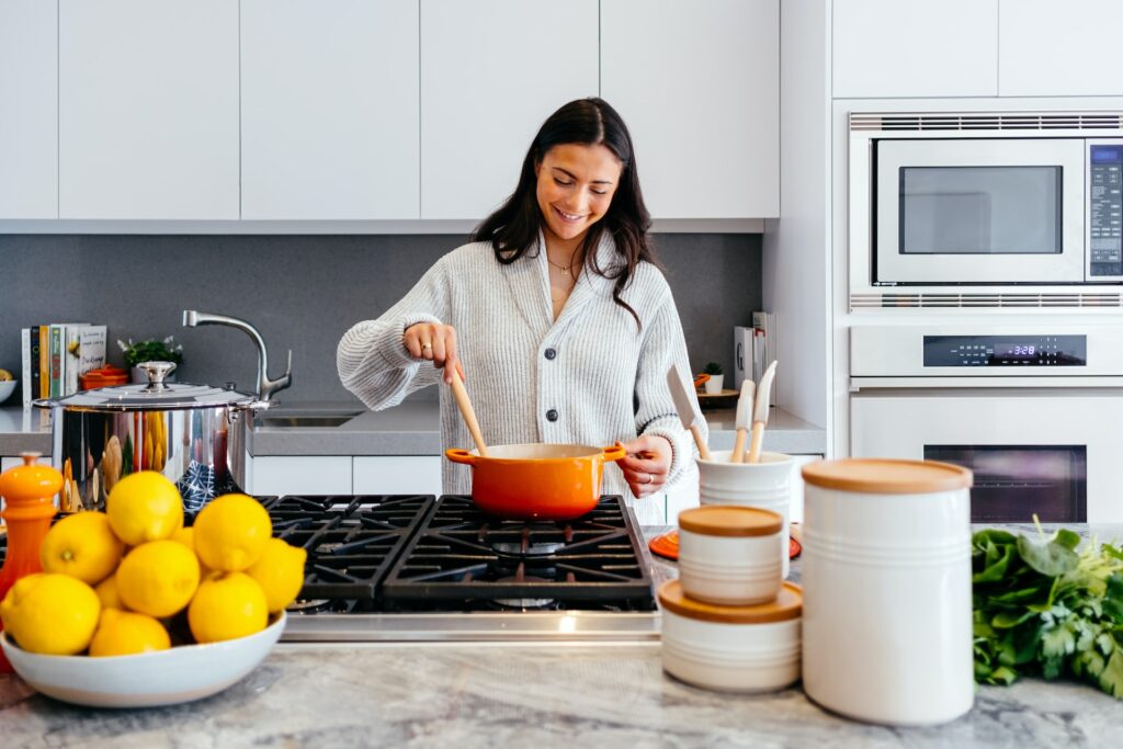 A woman wearing a gray sweater is holding a wooden ladle and an orange pot while cooking inside the kitchen