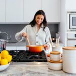 A woman wearing a gray sweater is holding a wooden ladle and an orange pot while cooking inside the kitchen