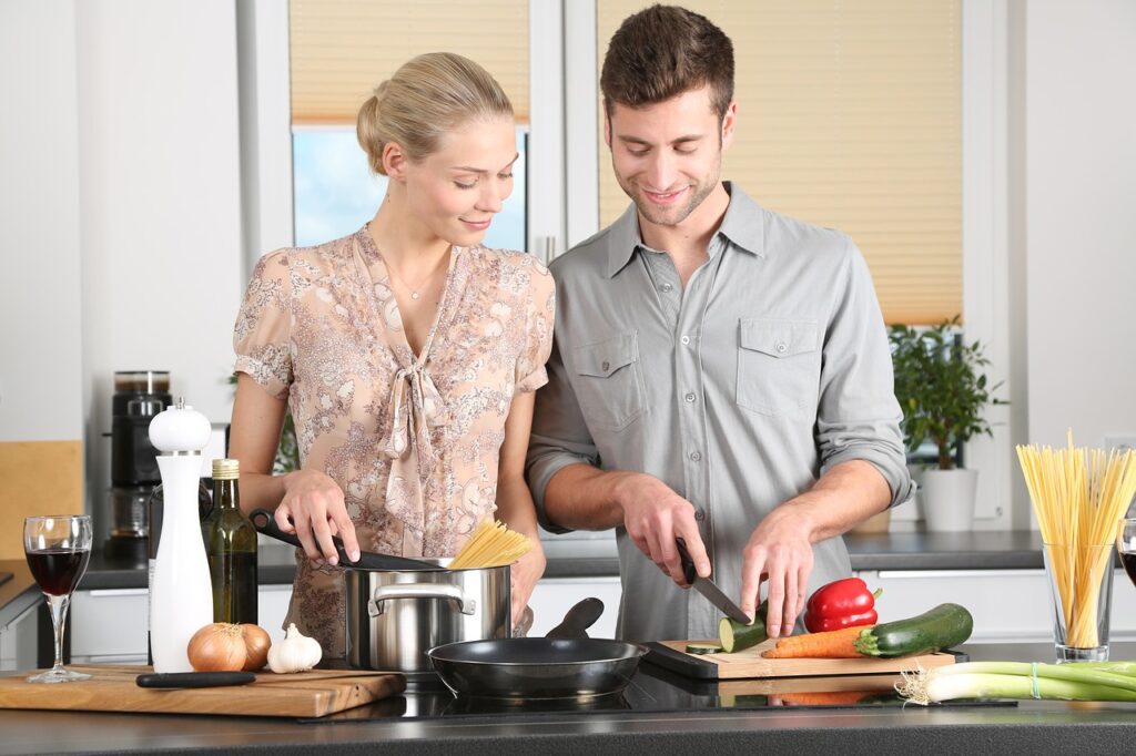 A woman wearing a floral dress is standing beside a man wearing a gray button shirt while slicing a fresh zucchini in the kitchen