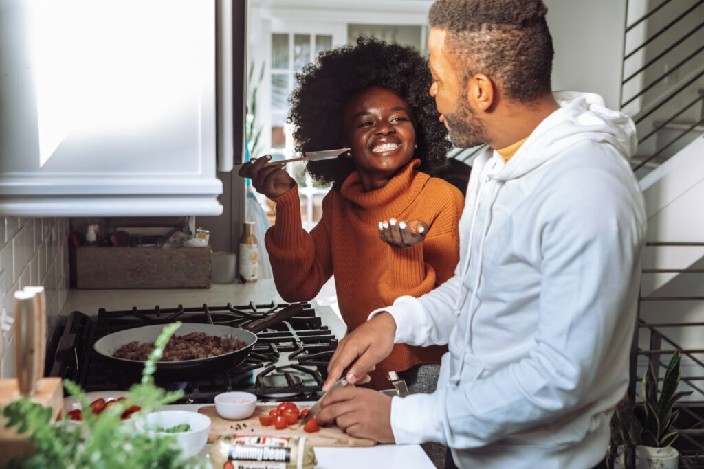 A woman wearing an orange turtleneck is standing beside a man wearing a gray hoodie near a black stovetop