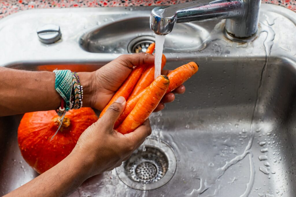 A person wearing colorful bracelets is washing fresh carrots on a stainless steel sink