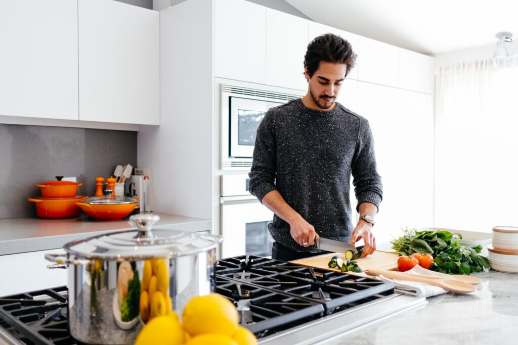 A man wearing a black sweatshirt is holding a stainless steel knife to cut a fresh cucumber on a wooden chopping board