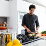 A man wearing a black sweatshirt is holding a stainless steel knife to cut a fresh cucumber on a wooden chopping board