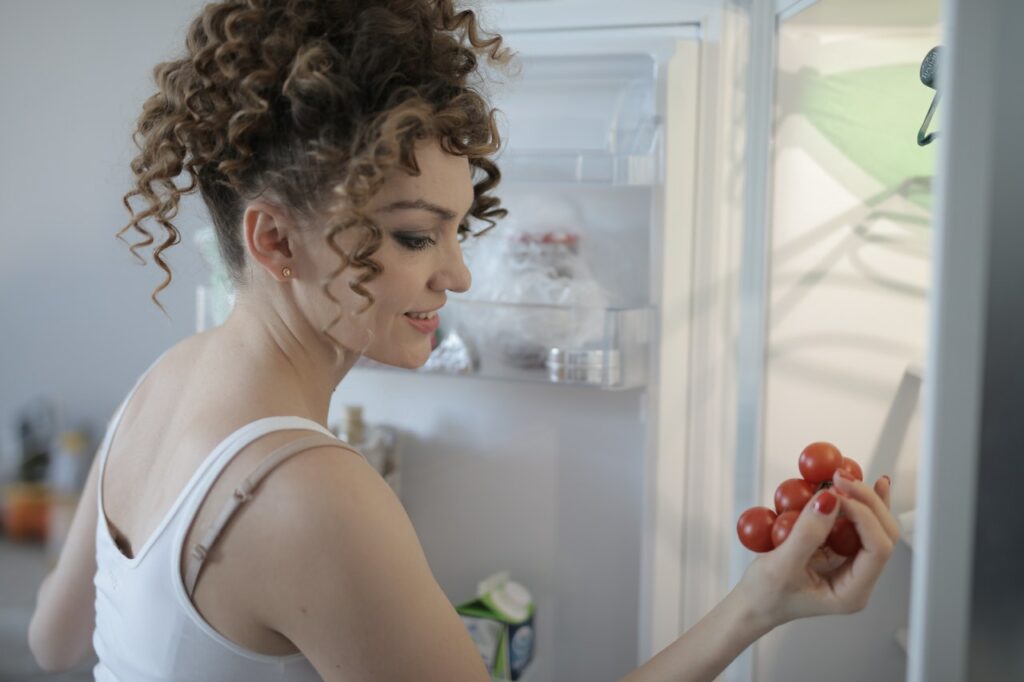 A woman with curly hair and wearing a white tank top is holding grapes near an open white fridge