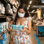 A woman wearing a floral dress and white face mask is holding a glass container inside a store