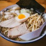 A ramen with a slice of egg on top and a white scoop placed on a white ceramic bowl