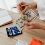 A person counting money while holding a gold pen near a blue calculator and notebook placed on top of a brown wooden table