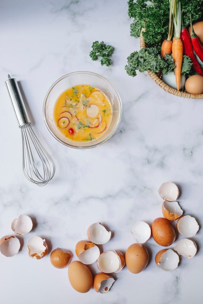 Whisked eggs mixed with sliced veggies placed on a clear glass bowl near a silver whisk, a basket of fresh vegetables, and egg shells on top of a marbled table
