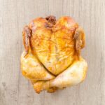 A roasted chicken near a silver and black knife on top of a brown wooden chopping board