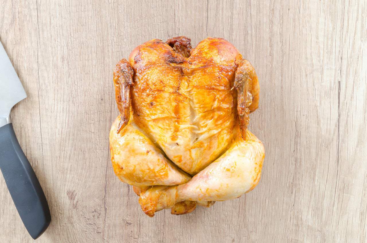A roasted chicken near a silver and black knife on top of a brown wooden chopping board