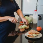 A woman wearing a navy blue dress is cutting lettuce on a brown wooden chopping board on top of a black countertop
