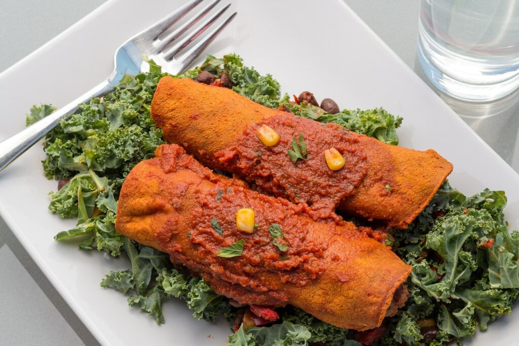 Two enchiladas with red sauce on top of green leafy vegetables on a white plate