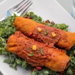 Two enchiladas with red sauce on top of green leafy vegetables on a white plate