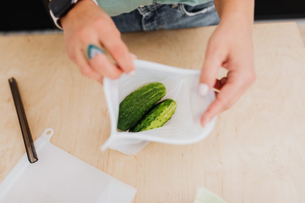 A person using a white resealable bag to store two cucumbers on top of a brown wooden table