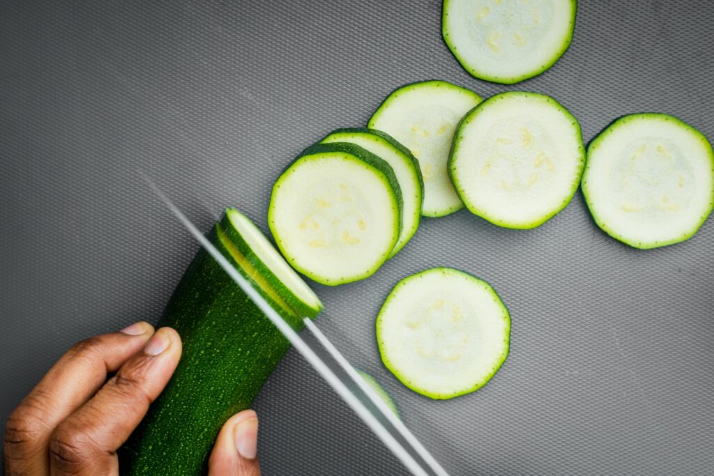 A person is using a stainless steel knife to cut thin slices of cucumber on a gray chopping board