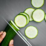A person is using a stainless steel knife to cut thin slices of cucumber on a gray chopping board