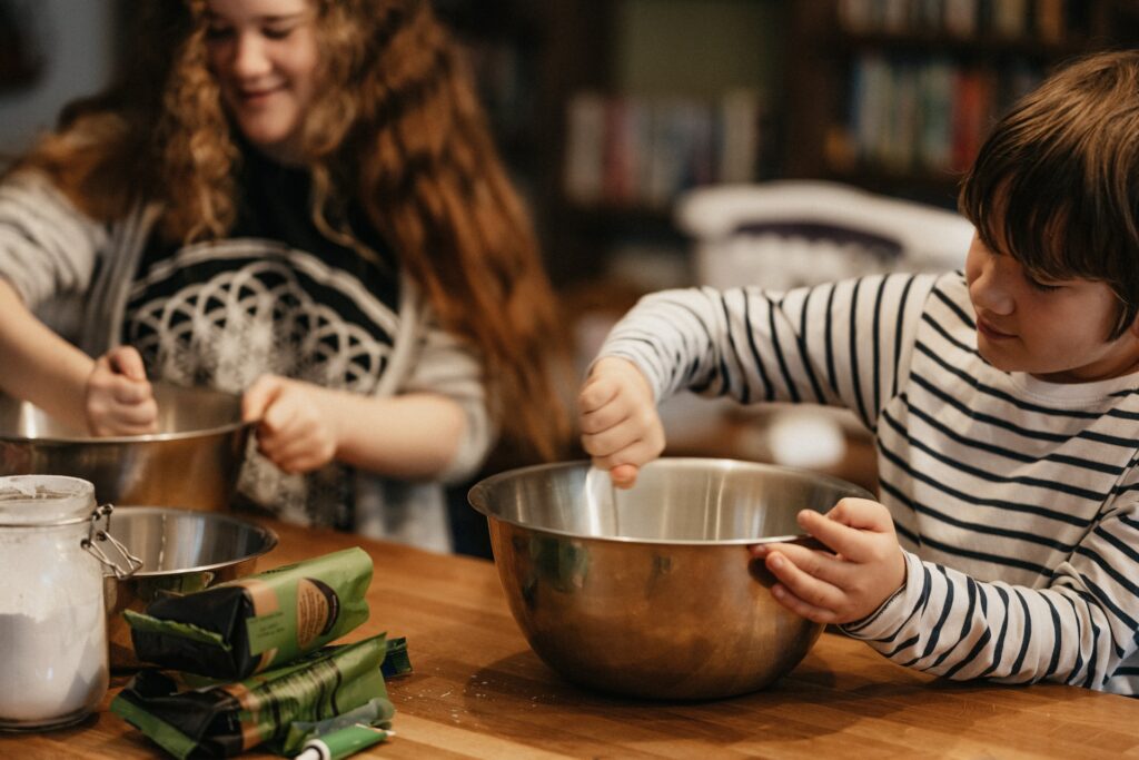 A woman with brown curly hair is mixing a stainless bowl next to a young boy mixing a bowl on top of a brown wooden table