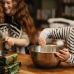 A woman with brown curly hair is mixing a stainless bowl next to a young boy mixing a bowl on top of a brown wooden table