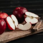 A sliced apple near three red apples and a knife on a wooden chopping board on top of a black wooden table