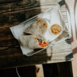 A burrito cut in half is placed on a white plate with a silver knife, and the burrito dip on top of a brown wooden table