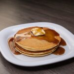 Pancakes with a slice of butter drizzled with syrup on a white ceramic plate on top of a brown wooden table