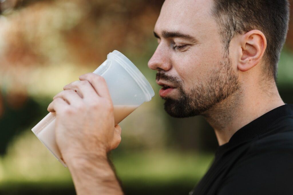 A man wearing a black shirt is holding a plastic tumbler with a brown protein shake