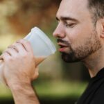 A man wearing a black shirt is holding a plastic tumbler with a brown protein shake