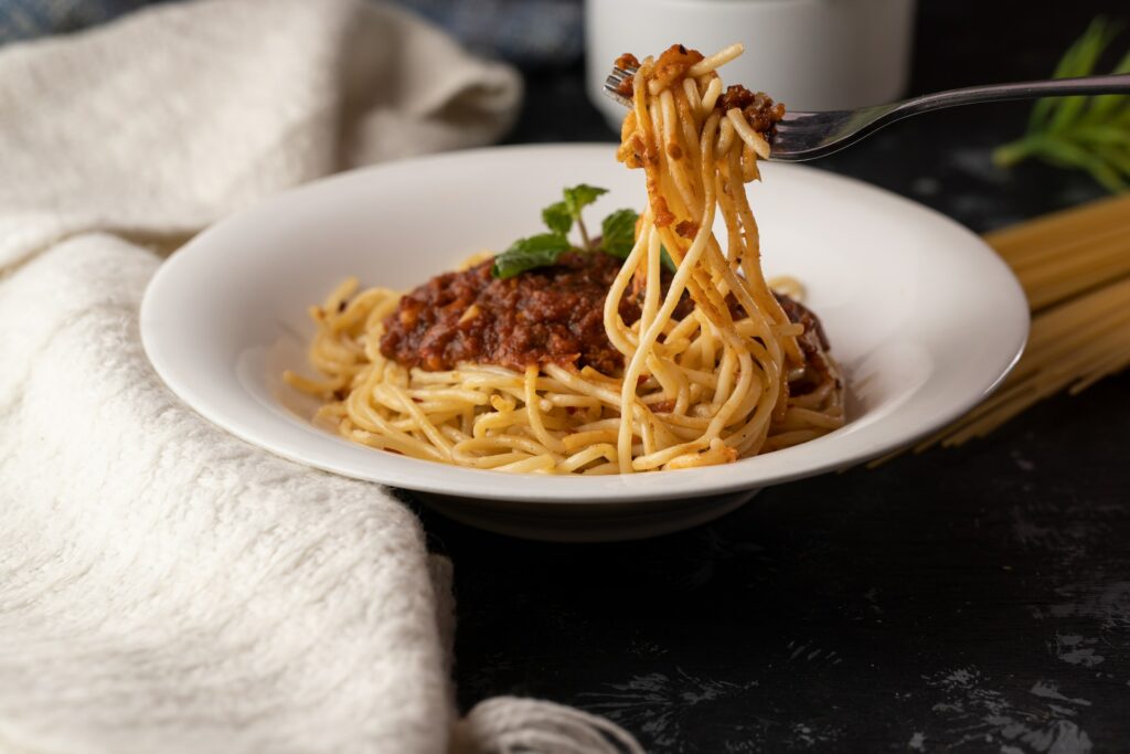A person using a silver fork to get spaghetti with fresh basil placed on a white ceramic bowl on top of a black table