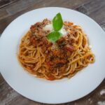 Spaghetti garnished with parmesan cheese and fresh basil on a white ceramic plate on top of a brown wooden table