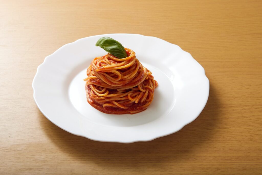 A person using a silver fork to get spaghetti with meatballs on a stainless steel frying pan on top of a gray surface