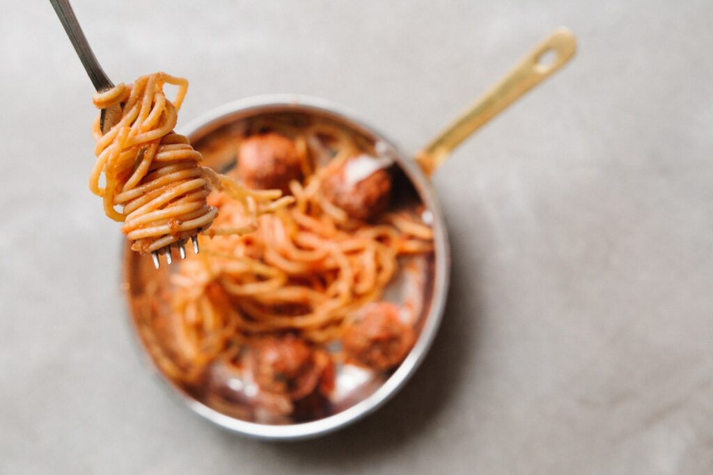 A person using a silver fork to get spaghetti with meatballs on a stainless steel frying pan on top of a gray surface