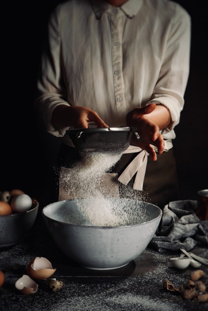 A baker sifting a flour on a strainer in a big white bowl
