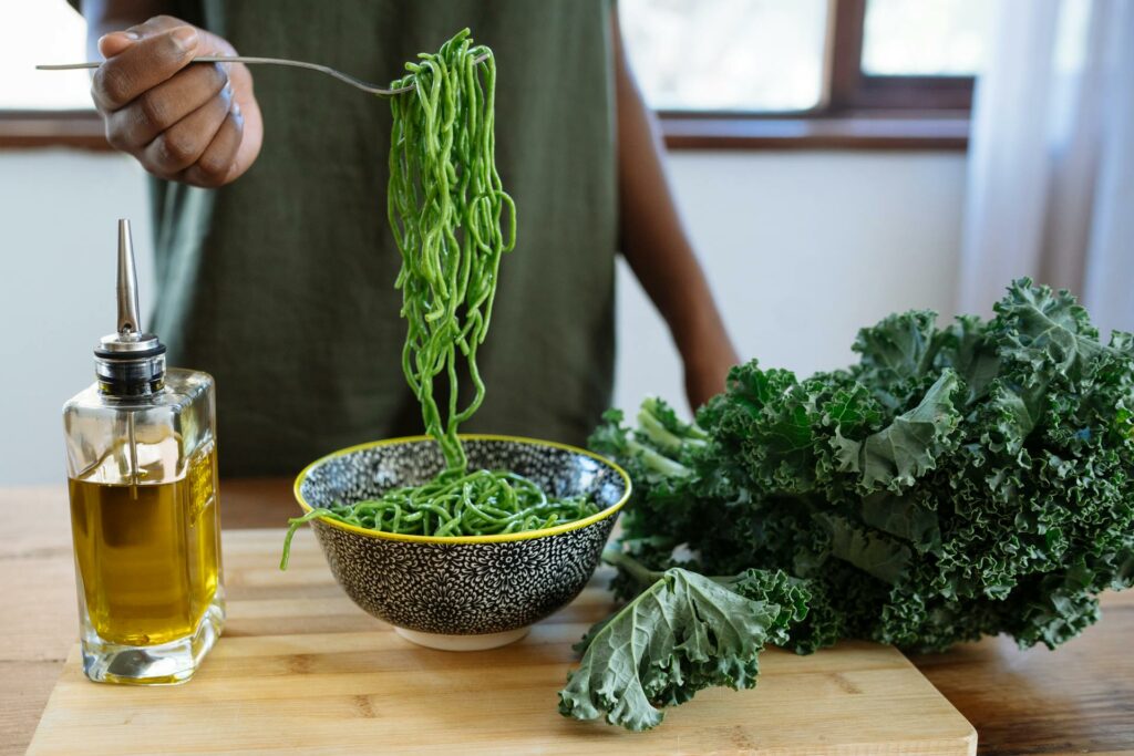 A woman holding a fork with green noodles beside a group of kale and oil